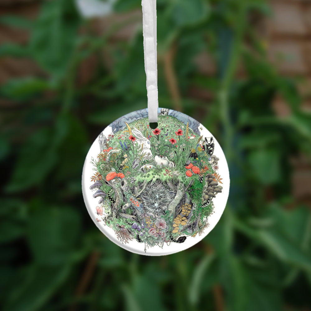 The other side of the ornament. A flower-filled meadow. A stag and badger stand either side, and a hare, blackbird, mouse, and other creatures are nestled within. The head and shoulders of a young woman lying on her back rise from the ground. Beneath her, the bottom half of the oval is a mass of twisting roots, branches, leaves, fungi and toadstools. A snake winds its way through the top half with its head appearing at the bottom.