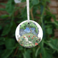 One side of the ornament. At the top there's a tree in full leaf, surrounded by clouds, but clearing to blue in the centre, and swifts wheeling all around. Beneath, the top half of the oval is a flower-filled meadow. A stag and badger stand either side, and a hare, blackbird, mouse, and other creatures are nestled within. The head and shoulders of a young woman lying on her back rise from the ground. 