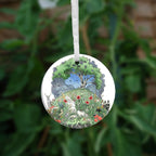 One side of the ornament. At the top there's a tree in full leaf, surrounded by clouds, but clearing to blue in the centre, and swifts wheeling all around. Beneath, the top half of the oval is a flower-filled meadow. A stag and badger stand either side, and a hare, blackbird, mouse, and other creatures are nestled within. The head and shoulders of a young woman lying on her back rise from the ground. 