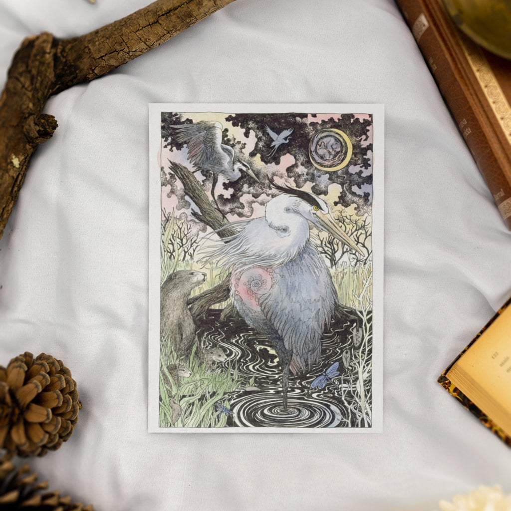 Greeting card of a heron in a natural setting on a textured surface with a pine cone and book.
