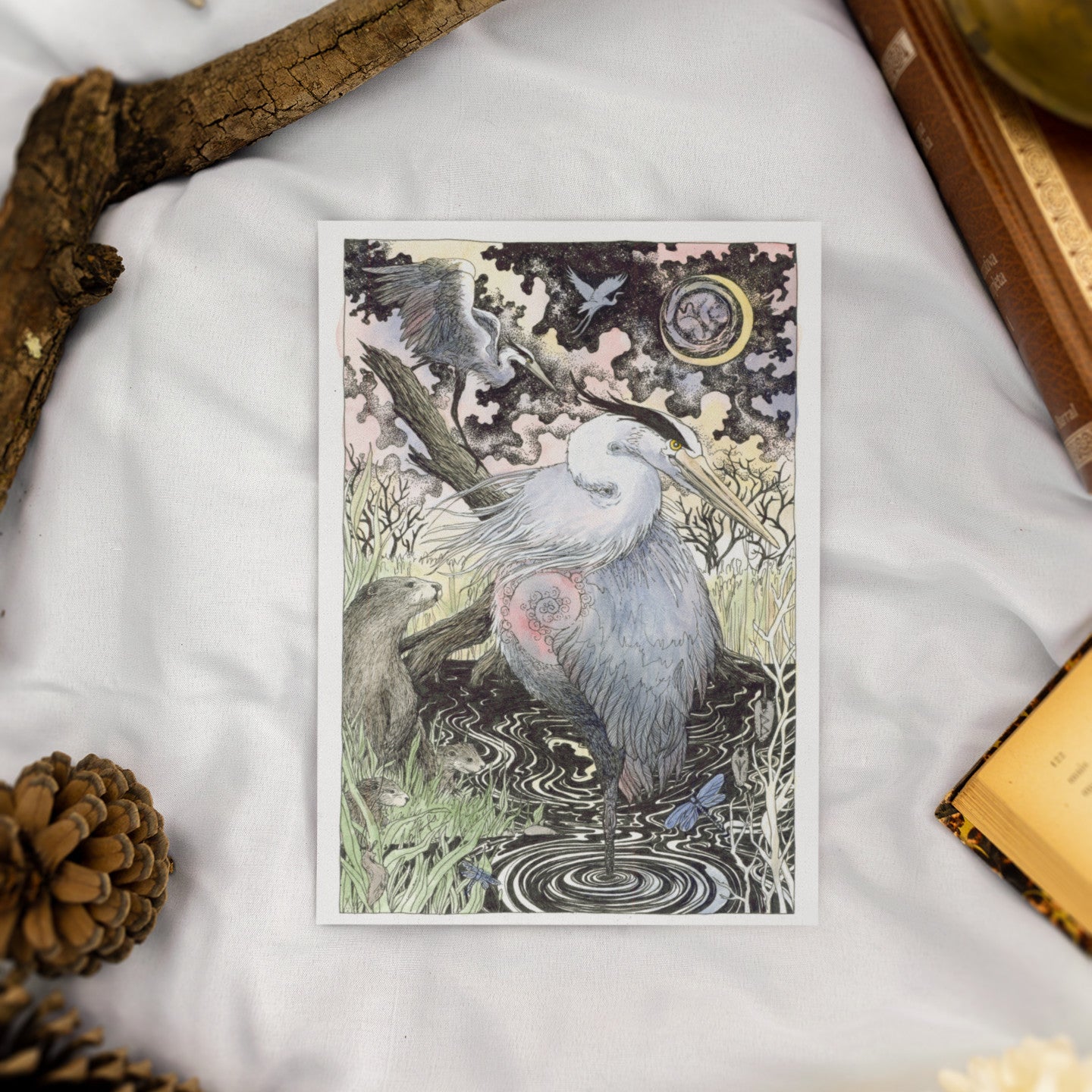 Greeting card of a heron in a natural setting on a textured surface with a pine cone and book.