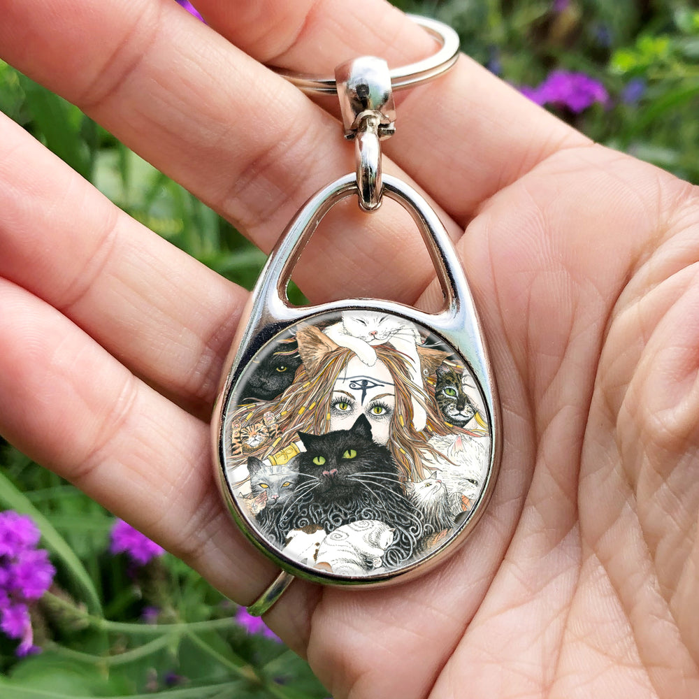 A mass of cats of all colours and in many poses surrounds the face of a red-haired woman with cat-slit eyes and ears, and a Eye of Horus symbol on her forehead. A white cat sits on her head. She holds a gold-coloured sistrum (a type of rattle) in one hand. Colours are oranges, reds, black and white.