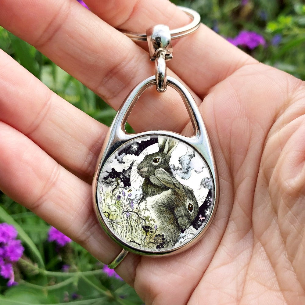 The head and body of two hares, one sitting upright facing to the left, the other, head down, facing right. They're in a meadow full of flowers, berries, butterflies, bees and dragonflies. Behind them a full moon rises surrounded by wispy, swirly clouds.