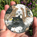 A mass of cats of all colours and in many poses surrounds the face of a red-haired woman with cat-slit eyes and ears, and a Eye of Horus symbol on her forehead. A white cat sits on her head. She holds a gold-coloured sistrum (a type of rattle) in one hand. Colours are oranges, reds, black and white.