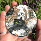A mass of cats of all colours and in many poses surrounds the face of a red-haired woman with cat-slit eyes and ears, and a Eye of Horus symbol on her forehead. A white cat sits on her head. She holds a gold-coloured sistrum (a type of rattle) in one hand. Colours are oranges, reds, black and white.