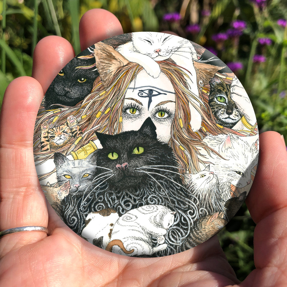 A mass of cats of all colours and in many poses surrounds the face of a red-haired woman with cat-slit eyes and ears, and a Eye of Horus symbol on her forehead. A white cat sits on her head. She holds a gold-coloured sistrum (a type of rattle) in one hand. Colours are oranges, reds, black and white.
