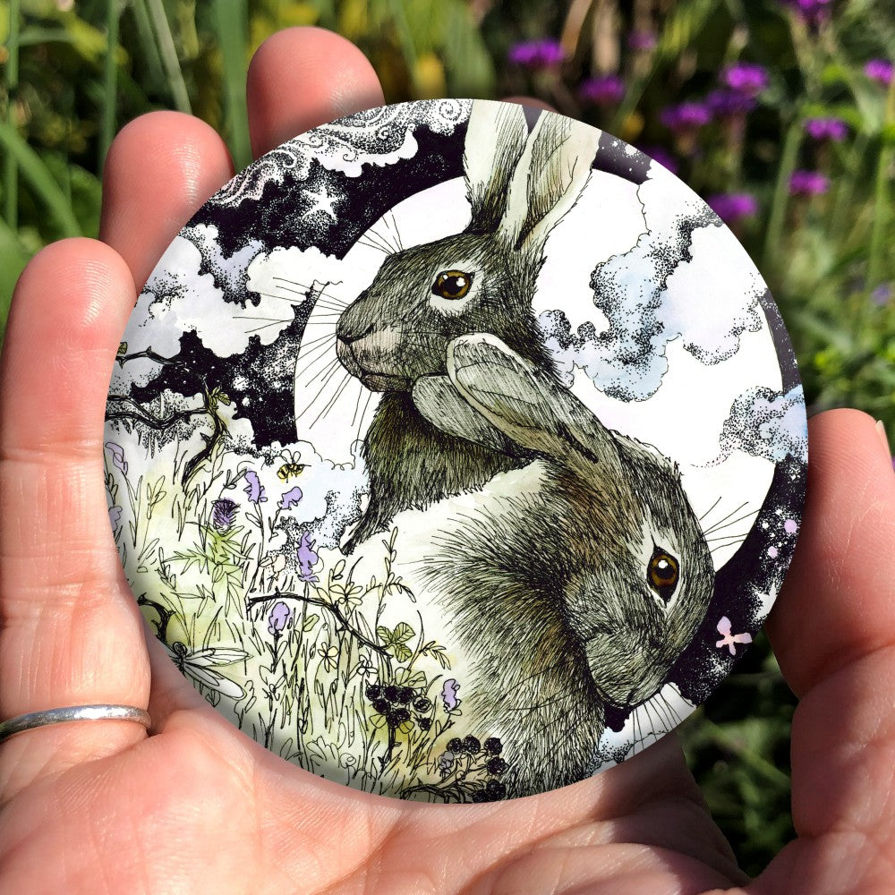 The head and body of two hares, one sitting upright facing to the left, the other, head down, facing right. They're in a meadow full of flowers, berries, butterflies, bees and dragonflies. Behind them a full moon rises surrounded by wispy, swirly clouds.