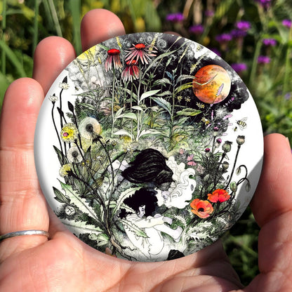 A figure in white with black hair is curled up in the foetal position. A second smaller figure in black with white hair lies above her. A multitude of plants and flowers surround them, rising upwards towards a sun and moon to the left and right, and red echinacea flowers in the centre.