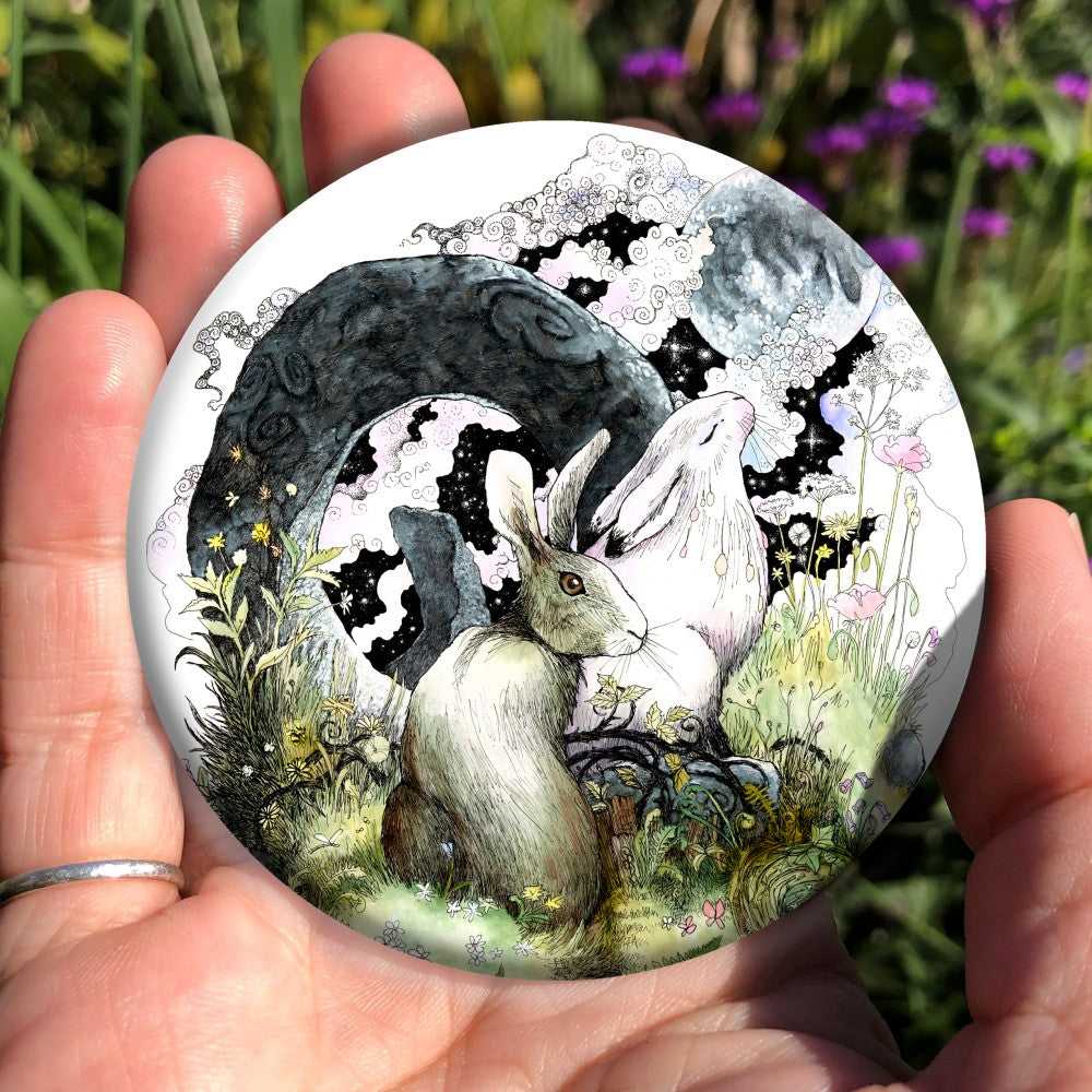 Two hares sit on grass in front of an upright grey circular stone with a large hole in the centre. one of the hares is turning to look towards the viewer, the other has their eyes closed and head tilted upward. 