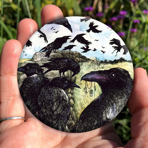 A large flock of ravens sit on and fly around a drystone wall surrounded by yellow-green grassy fields. The blue sky is broken with white clouds. In the foreground a raven holds a string of tokens in its beak, including a pentagram, key and Teiwaz rune.