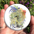 A cockerel with swirling blue, green and yellow/orange feathers, also incorporating leaves and wheat ears. Mice nibble on the ears, and butterflies and dragonflies flutter and swoop around. At the bottom, a crescent moon peeks from behind sprigs of berries. A yellow sun appears behind the head of the cockerel. A corn dolly is formed in the centre of the swirl of feathers, and a mouse sits in its skirt.