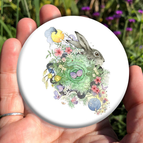 Two grey/blue eggs sit within a spiral with a green background. The spiral is surrounded by spring flowers, and a snake eating its tail weaves through them. The head of a hare or rabbit appears at the top right. A yellow circle surrounded by storm clouds appears above, and a blue disc surrounded by yellow flowers appears below.