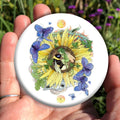 A bee and fairy curl up asleep in the centre of a sunflower. Blue butterflies flutter around them. Ivy and oak twine through the petals. The heads of two dragons can be seen above and below, behind the sunflower. A yellow/orange spiralled disc sits at the top, and similar disc with a star sits at the bottom.