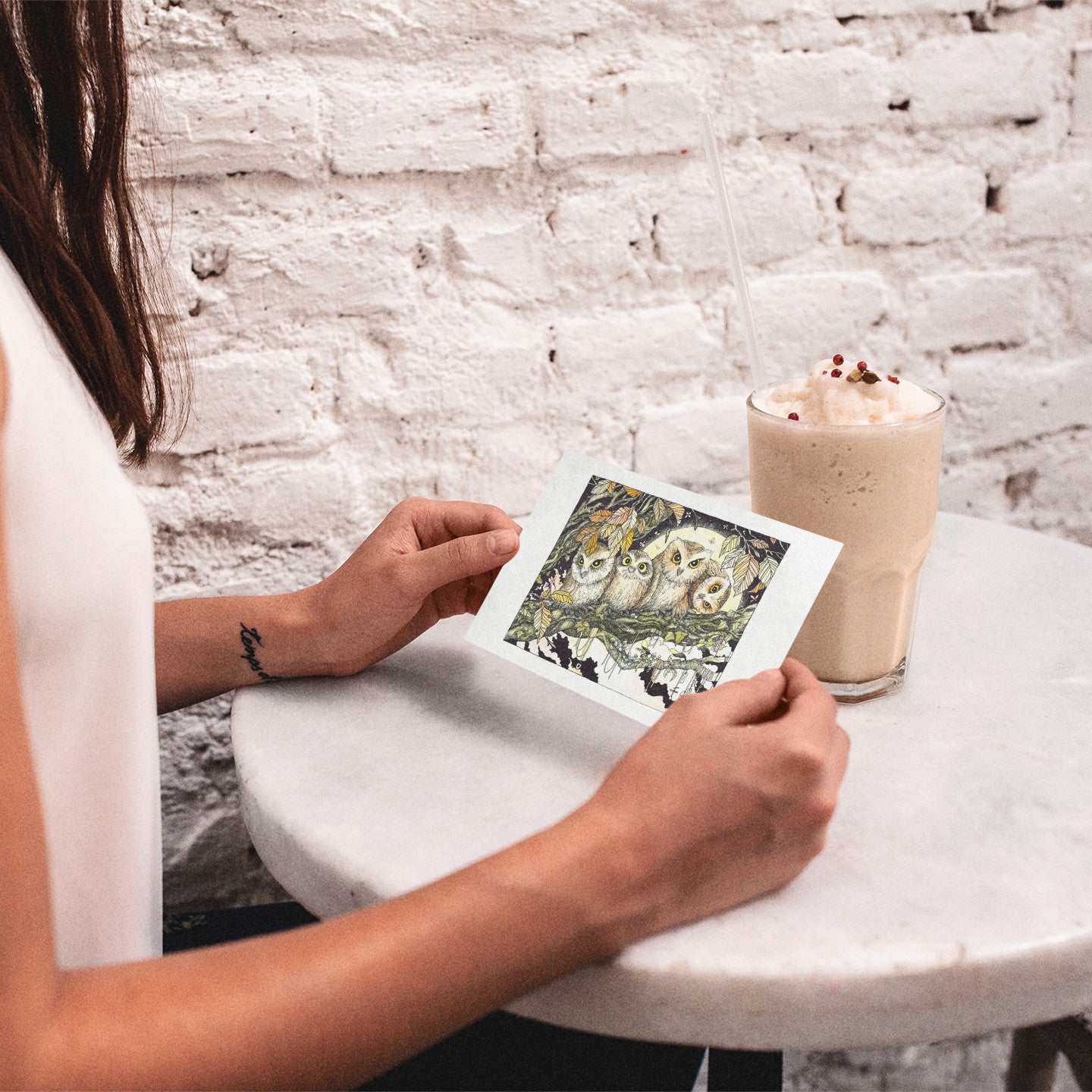Person holding a card with owl illustration next to a milkshake on a white table against a white brick wall.