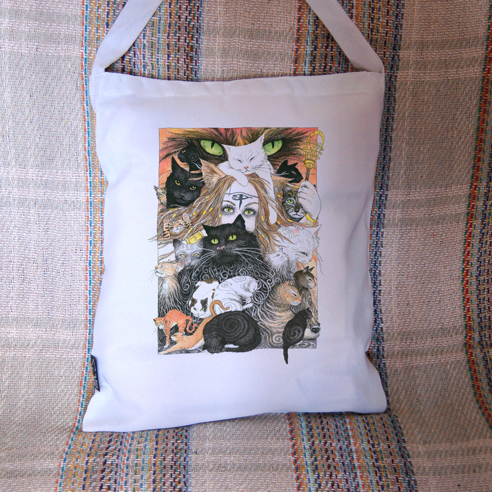 A mass of cats of all colours and in many poses surrounds the face of a red-haired woman with cat-slit eyes and ears, and a Eye of Horus symbol on her forehead. A white cat sits on her head. She holds a gold-coloured sistrum (a type of rattle) in one hand. The head of a much larger cat with green eyes appears above and behind them. Colours are oranges, reds, black and white.