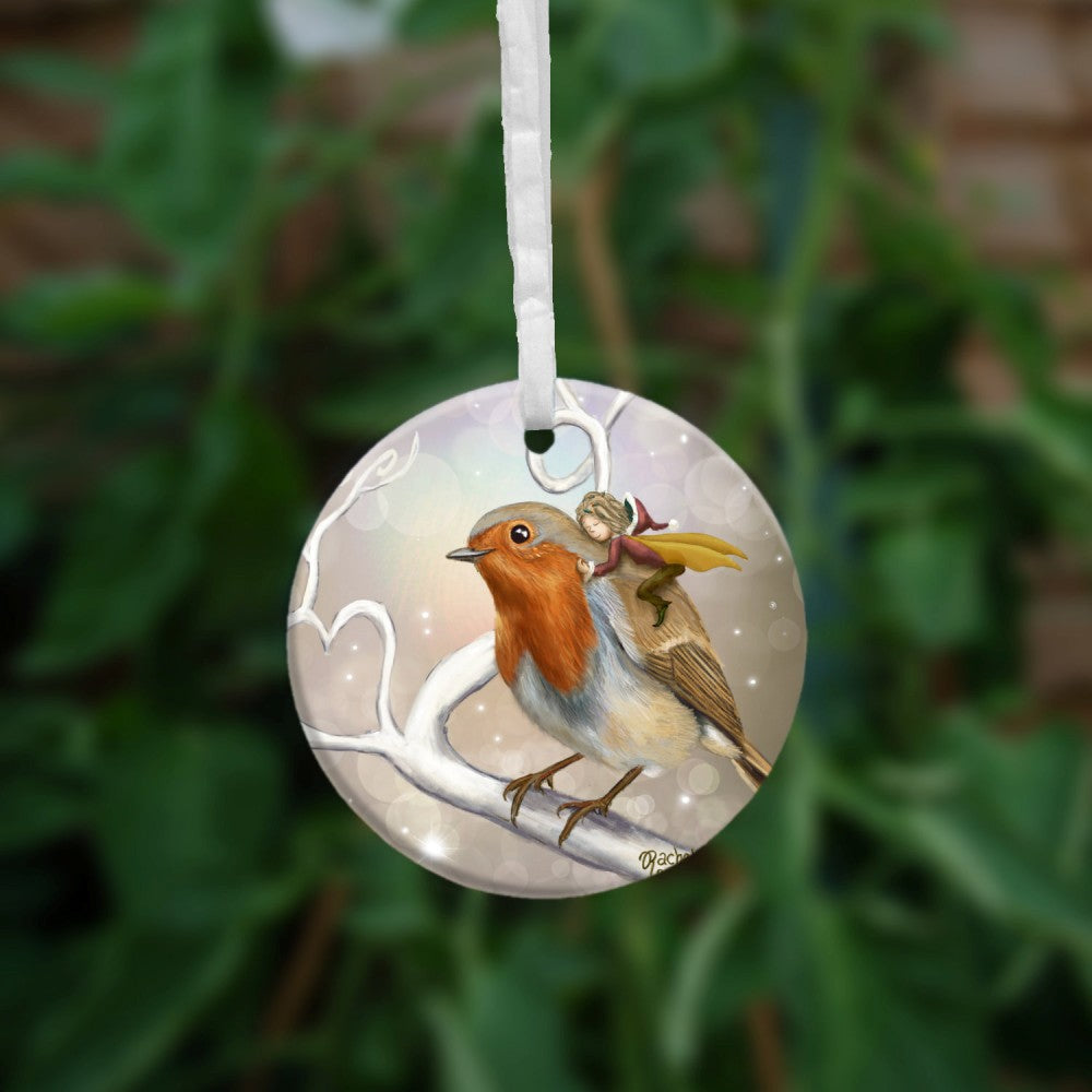 A robin perches on a white leafless branch. Twigs twist and curl from the branch, one forming a heart shape. A fairy with mustard-coloured wings and wearing red and brown, and a santa hat, holds onto the back of the robin, eyes closed and looking content. The background is mid-brown with stars and bubbles. 