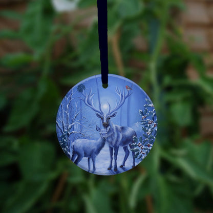 A stag and doe stand facing each other, heads turned towards the viewer. The design is cast in shades of blue. A robin perches on one of the stag's antlers. Snow lies underfoot, and on the branches of the berried holly tree to the right, and bare tree to the left. A wintery moon rises from between the stag's antlers, and stars can be seen in the sky beyond. 