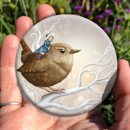 A smiling fairy in a mid-blue coat and hat and purple boots sits on the back of a wren. The wren sits on a white branch. Apart from the fairy, colours are browns and whites.