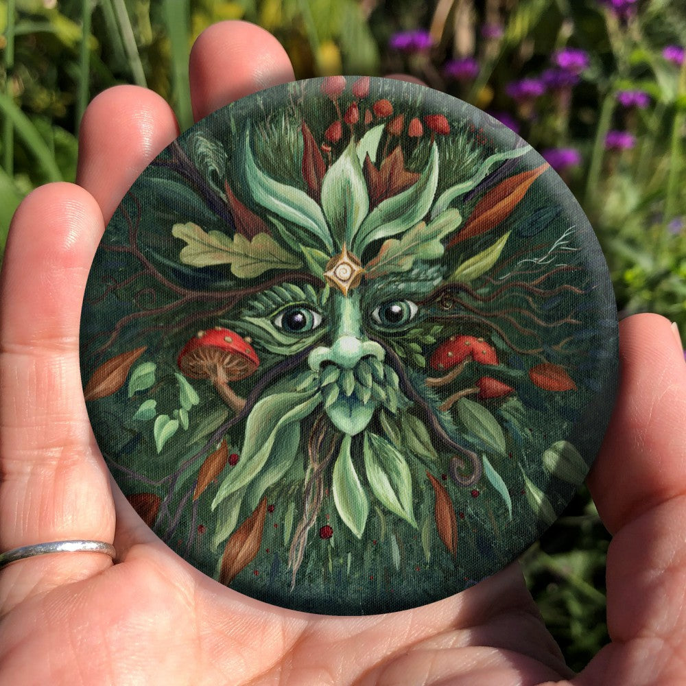 A pair of green eyes, with prominent nose beneath, is surrounded by red and green leaves radiating out from the central face. Red toadstools grow out from his cheeks and the top of his head. Two delicate tree branches protrude like antlers. The background is dark green.