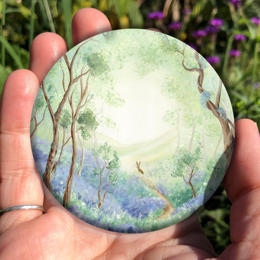 Trees reach upwards either side of a narrow winding path in woodland, the ground beneath them covered in bluebells. The path ends in the mid-distance, where a hare is sat. Beyond the hare, the trees open out to show hills in the distance. Colours are greens, blues and browns.