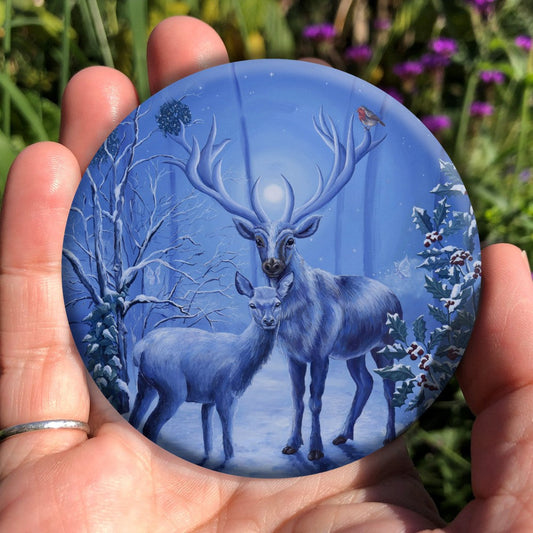 A stag and doe stand facing each other, heads turned towards the viewer. The design is cast in shades of blue. A robin perches on one of the stag's antlers. Snow lies underfoot, and on the branches of the berried holly tree to the right, and bare tree to the left. Pale fairies flit through the branches. Balls of mistletoe sit in the bare tree. A wintery moon rises from between the stag's antlers, and stars can be seen in the sky beyond. 