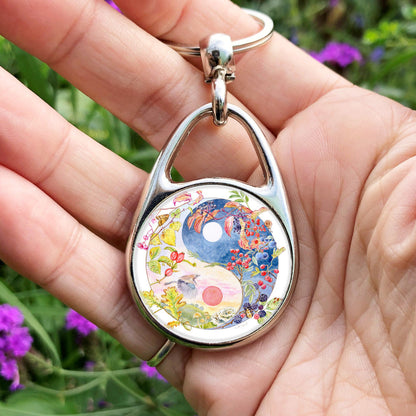 A Yin-Yang shape is surrounded by branches of native trees in autumn colours and some bearing fruit. The left hand half of the shape is filled with a pink/pale orange sky, and a large red sun forms the traditional 'dot' over a grassy hill. Autumn leaves and berries overlay the sky, a jay sings flies over, and fungi sit at the bottom. A moth sits on the outside curve. The right hand half features a dark blue sky with stars, with a white moon forming the 'dot'. Autumn leaves and berries overlay the scene.