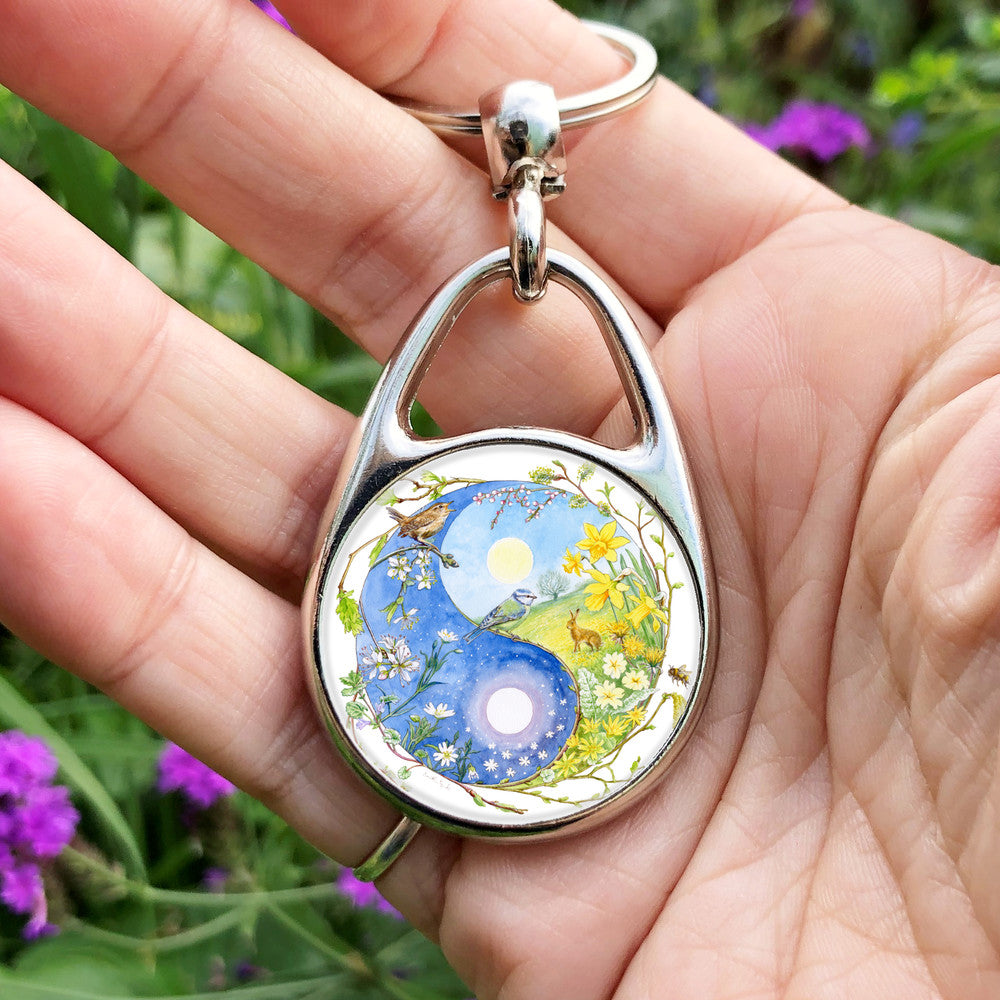 A Yin-Yang shape is surrounded by sprouting branches of native trees. The left hand half of the shape is filled with blue sky, and a large, watery sun forms the traditional 'dot'. White blossom and spring flowers overlay the sky, and a wren sings from a branch at the top. The right hand half features a hare sitting in a grassy field in a sunny day. A pale yellow sun forms the 'dot'. Yellow flowers overlay the scene. A blue tit sits on a branch forming the curve between the two sections.