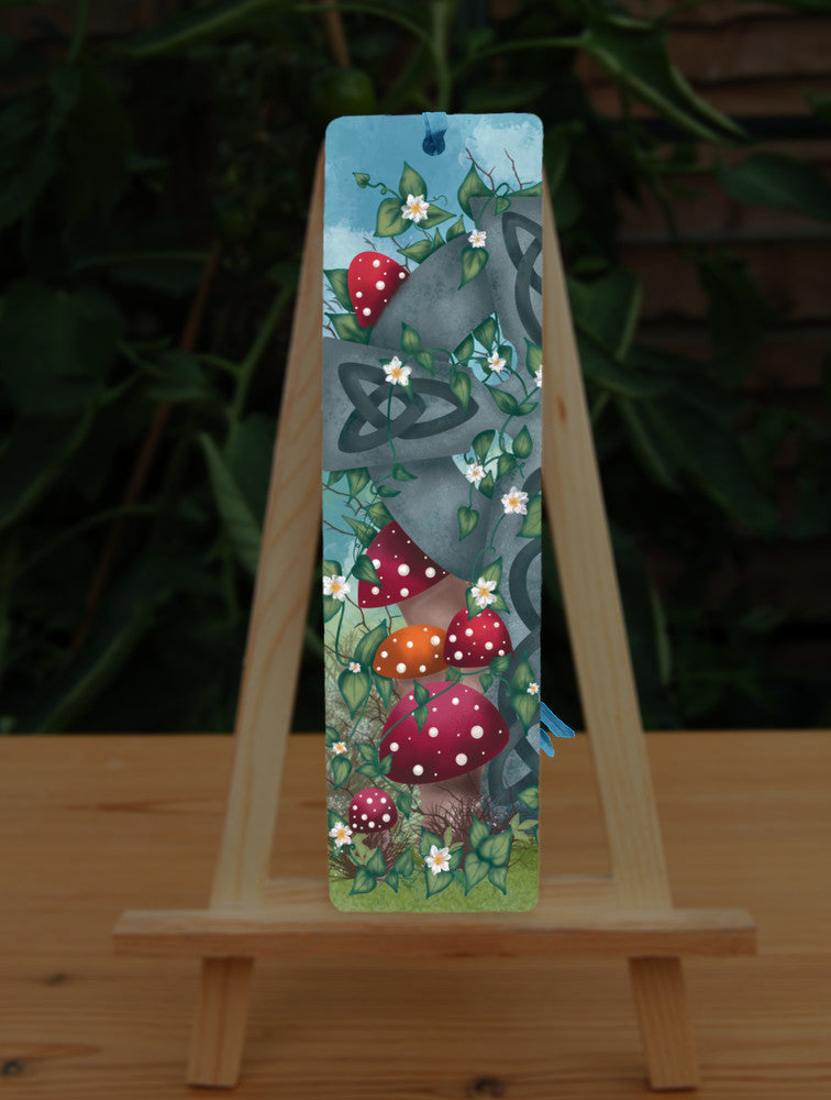 One half of a grey stone Celtic cross, inlaid with Celtic knots, stands in long grass surrounded by red and orange spotted toadstools. Leaves and small white flowers twine upwards and through the cross, and more toadstools appear near the top. There's a mottled blue sky in the background.