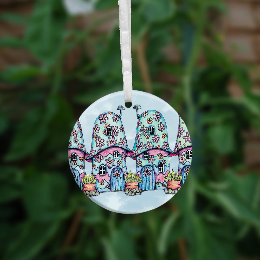 Four pink and blue / green toadstool houses stand in a row. The roofs and walls of all four are liberally decorated with simple flower designs have curved windows (two stories, plus a window in the roof) and have curved blue front doors. Tiny toadstool-shaped chimney pots protrude from each roof, and red planters surrounded by stones and with leafy plants growing from each sit between each house. The background is a mottled pale blue.