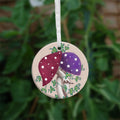 Two toadstools with long pale stems are twined together. The cap of one of them is red with white spots, the other purple with white spots. Leafy green tendrils twine around and up the pair. The background is a mottled beige colour.