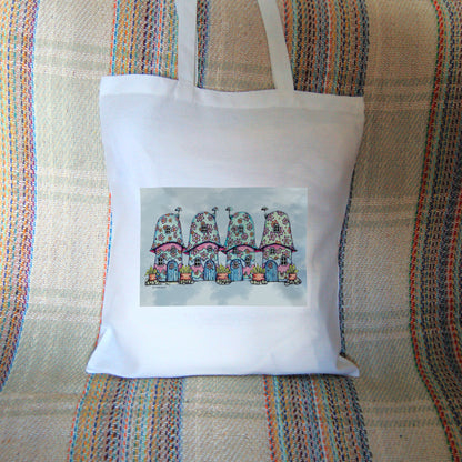 Four pink and blue / green toadstool houses stand in a row. The roofs and walls of all four are liberally decorated with simple flower designs have curved windows (two stories, plus a window in the roof) and have curved blue front doors. Tiny toadstool-shaped chimney pots protrude from each roof, and red planters surrounded by stones and with leafy plants growing from each sit between each house. The background is a mottled pale blue.