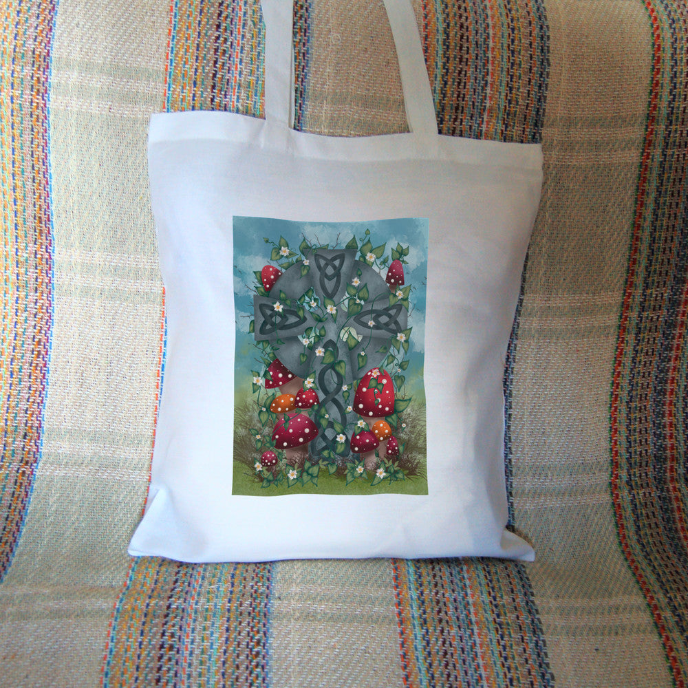 A grey stone Celtic cross, inlaid with Celtic knots, stands in long grass surrounded by red and orange spotted toadstools. Leaves and small white flowers twine upwards and through the cross, and more toadstools appear near the top. There's a mottled blue sky in the background.