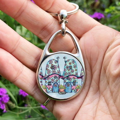 Two pink and blue / green toadstool houses stand in a row. The roofs and walls are liberally decorated with simple flower designs have curved windows (two stories, plus a window in the roof) and have curved blue front doors. Tiny toadstool-shaped chimney pots protrude from each roof, and red planters surrounded by stones and with leafy plants growing from each sit between each house. The background is a mottled pale blue.