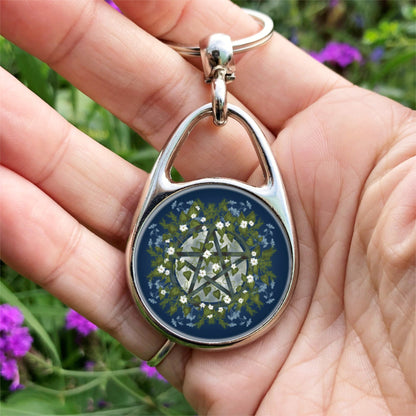 A pentacle symbol overlaid with green leaves, foliage and white flowers which extend outwards towards the edge of the design. A silvery full moon shines behind and within the circle of the pentacle. The background is deep blue.