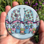 Two pink and blue / green toadstool houses stand in a row. The roofs and walls are liberally decorated with simple flower designs have curved windows (two stories, plus a window in the roof) and have curved blue front doors. Tiny toadstool-shaped chimney pots protrude from each roof, and red planters surrounded by stones and with leafy plants growing from each sit between each house. The background is a mottled pale blue.