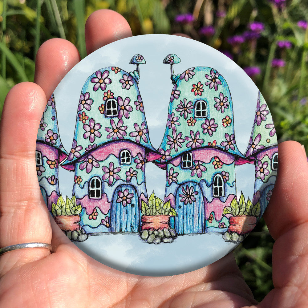 Two pink and blue / green toadstool houses stand in a row. The roofs and walls are liberally decorated with simple flower designs have curved windows (two stories, plus a window in the roof) and have curved blue front doors. Tiny toadstool-shaped chimney pots protrude from each roof, and red planters surrounded by stones and with leafy plants growing from each sit between each house. The background is a mottled pale blue.
