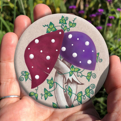 Two toadstools with long pale stems are twined together. The cap of one of them is red with white spots, the other purple with white spots. Leafy green tendrils twine around and up the pair. The background is a mottled beige colour.