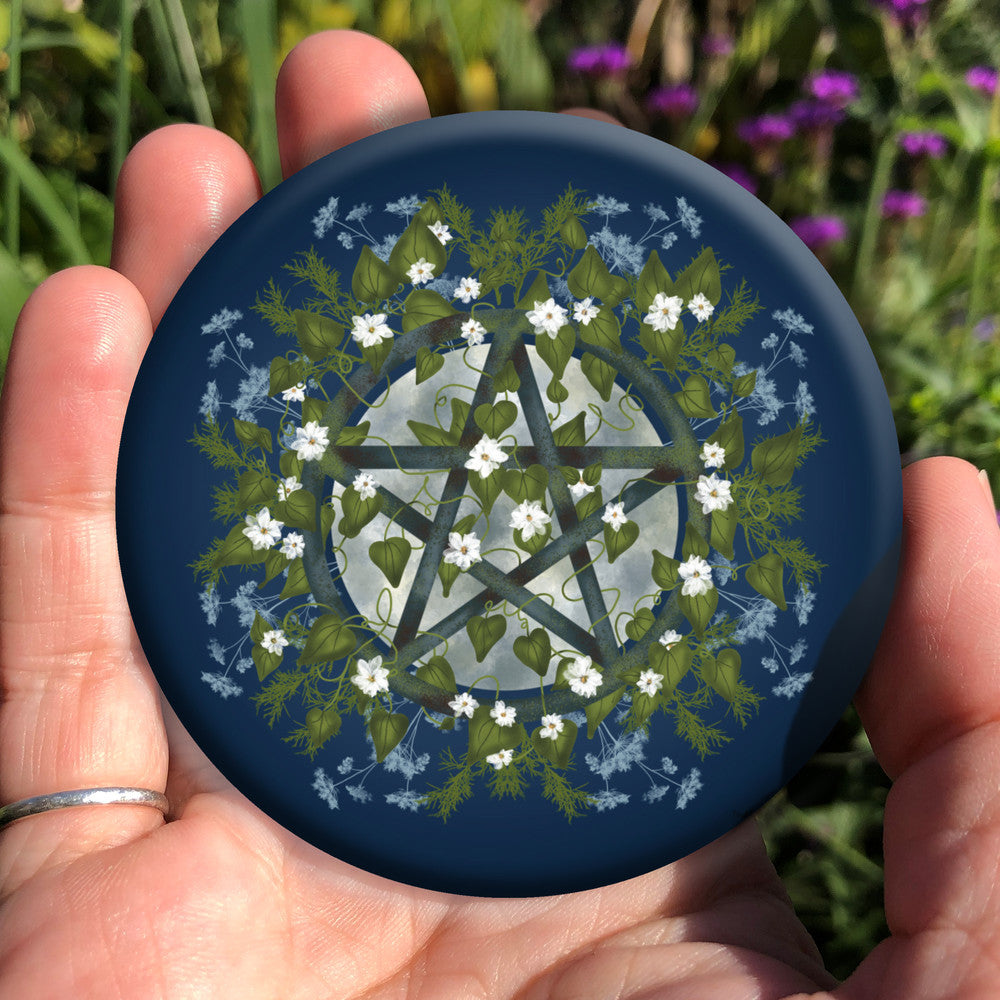 A pentacle symbol overlaid with green leaves, foliage and white flowers which extend outwards towards the edge of the design. A silvery full moon shines behind and within the circle of the pentacle. The background is deep blue.