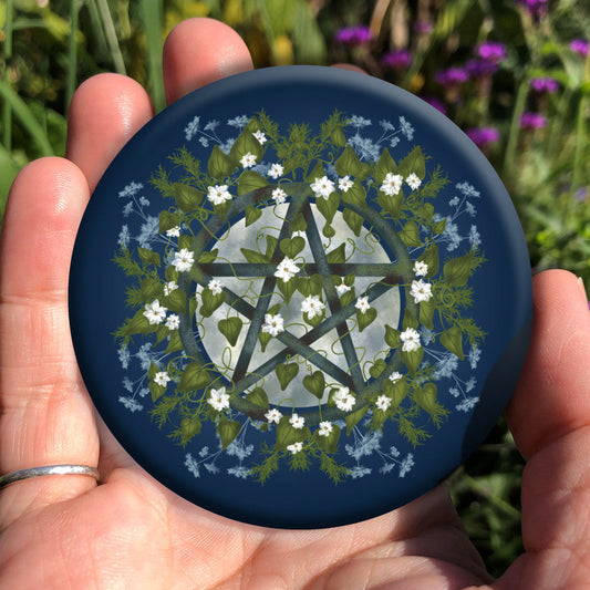 A pentacle symbol overlaid with green leaves, foliage and white flowers which extend outwards towards the edge of the design. A silvery full moon shines behind and within the circle of the pentacle. The background is deep blue.