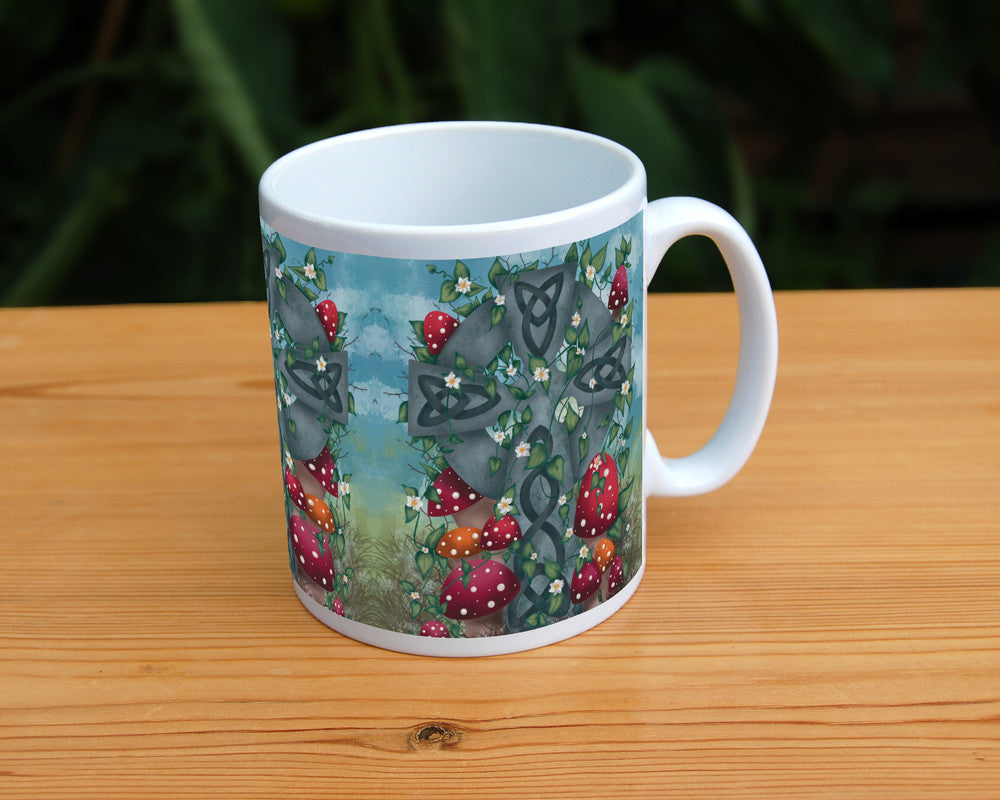 A grey stone Celtic cross, inlaid with Celtic knots, stands in long grass surrounded by red and orange spotted toadstools. Leaves and small white flowers twine upwards and through the cross, and more toadstools appear near the top. There's a mottled blue sky in the background.