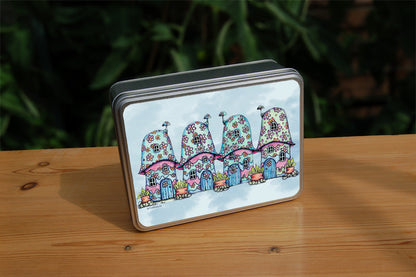 Four pink and blue / green toadstool houses stand in a row. The roofs and walls of all four are liberally decorated with simple flower designs have curved windows (two stories, plus a window in the roof) and have curved blue front doors. Tiny toadstool-shaped chimney pots protrude from each roof, and red planters surrounded by stones and with leafy plants growing from each sit between each house. The background is a mottled pale blue.